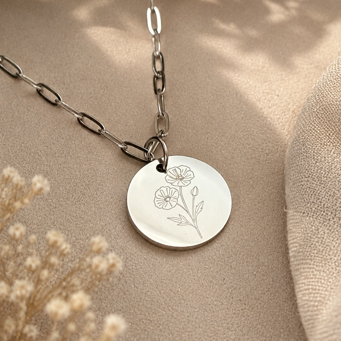 A silver disc pendant necklace with an engraved floral design, hanging from a silver link chain on a beige textured surface with dried flowers and green leaves in the background.