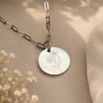 A silver disc pendant necklace with an engraved floral design, hanging from a silver link chain on a beige textured surface with dried flowers and green leaves in the background.