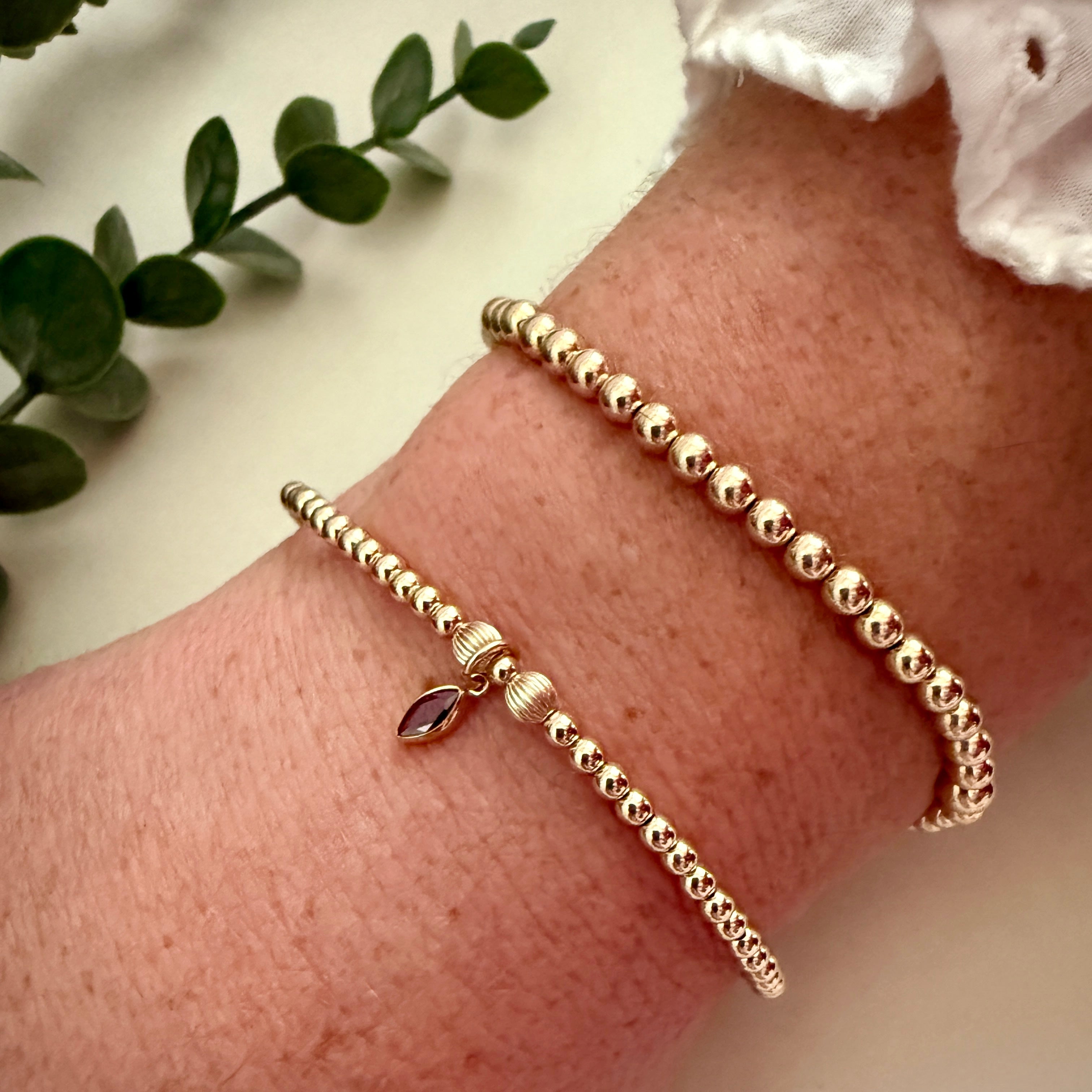 Gold beaded bracelet with a purple gemstone on a wrist, with a light background and green leaves.