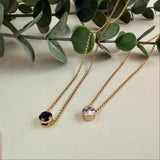 Two gold necklaces with gemstone pendants on a light surface with green leaves in the background