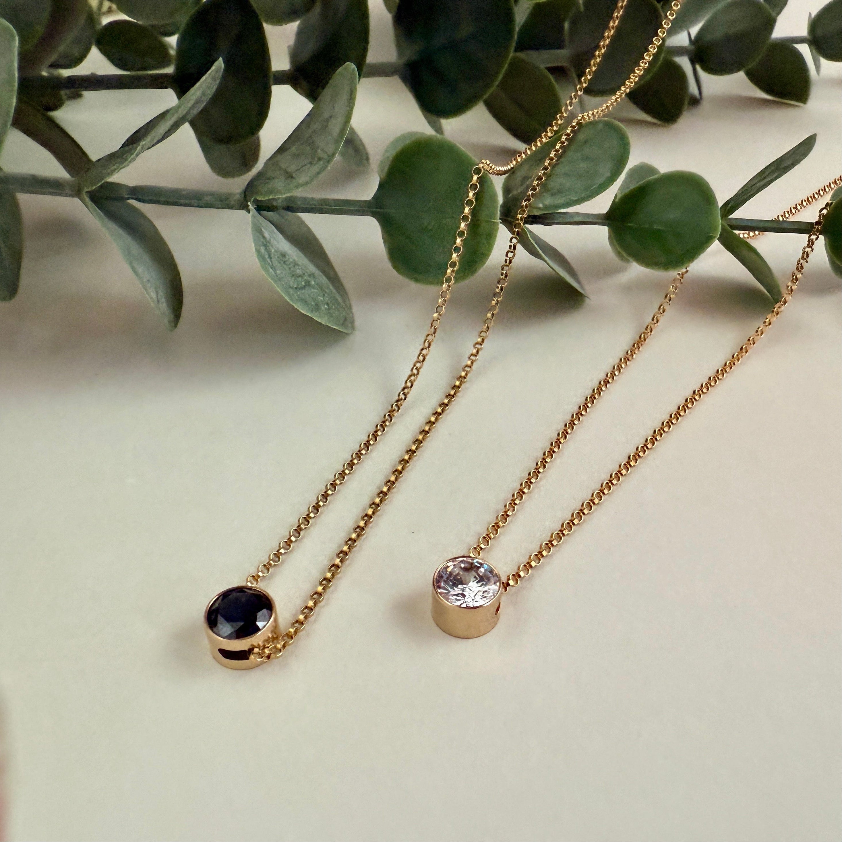 Two gold necklaces with gemstone pendants on a light surface with green leaves in the background