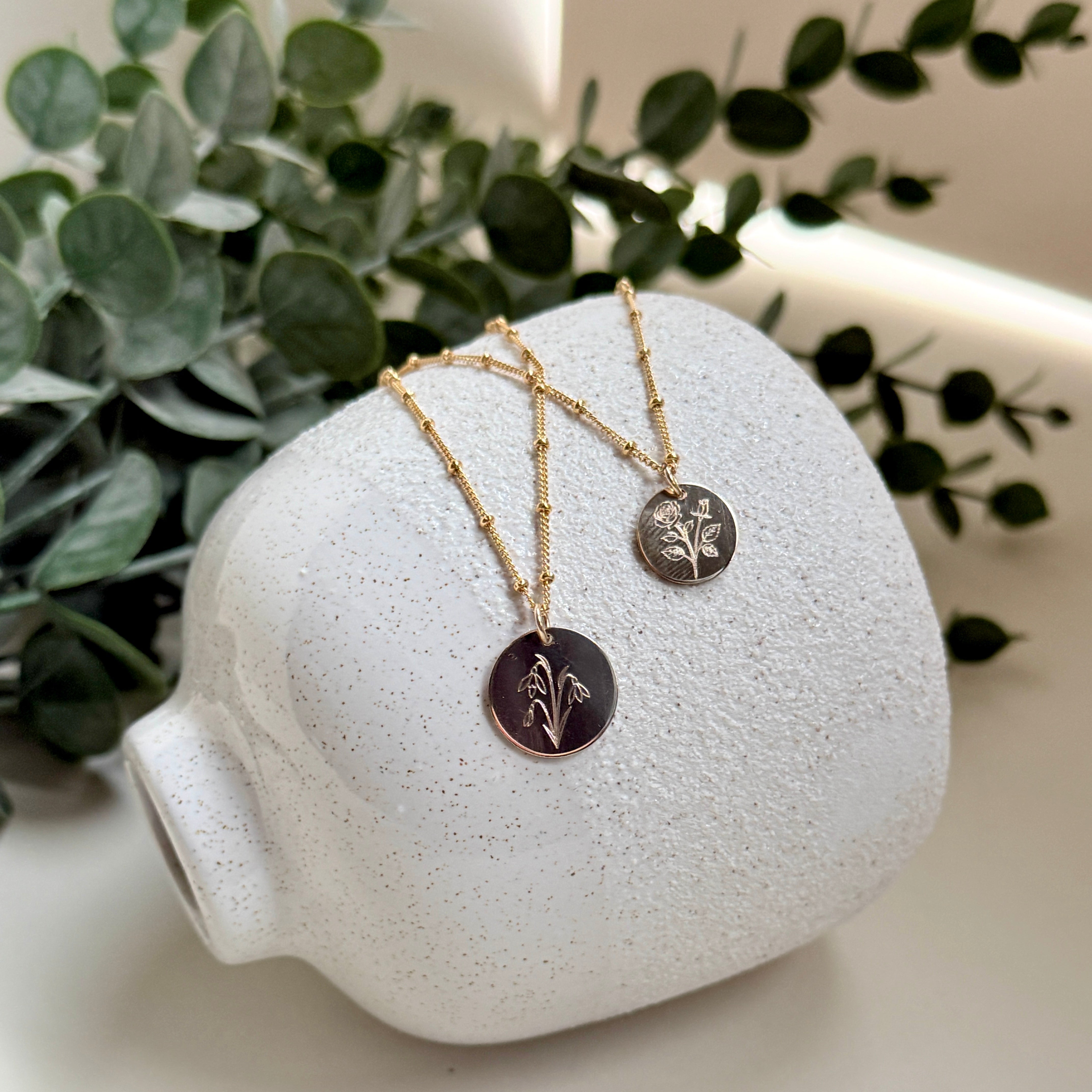 Two necklaces with circular pendants on a white ceramic stand with green leaves in the background