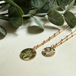 Two gold necklaces with circular pendants on a light surface with green leaves in the background