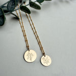 Two gold necklaces with engraved designs on a gray surface with green leaves.