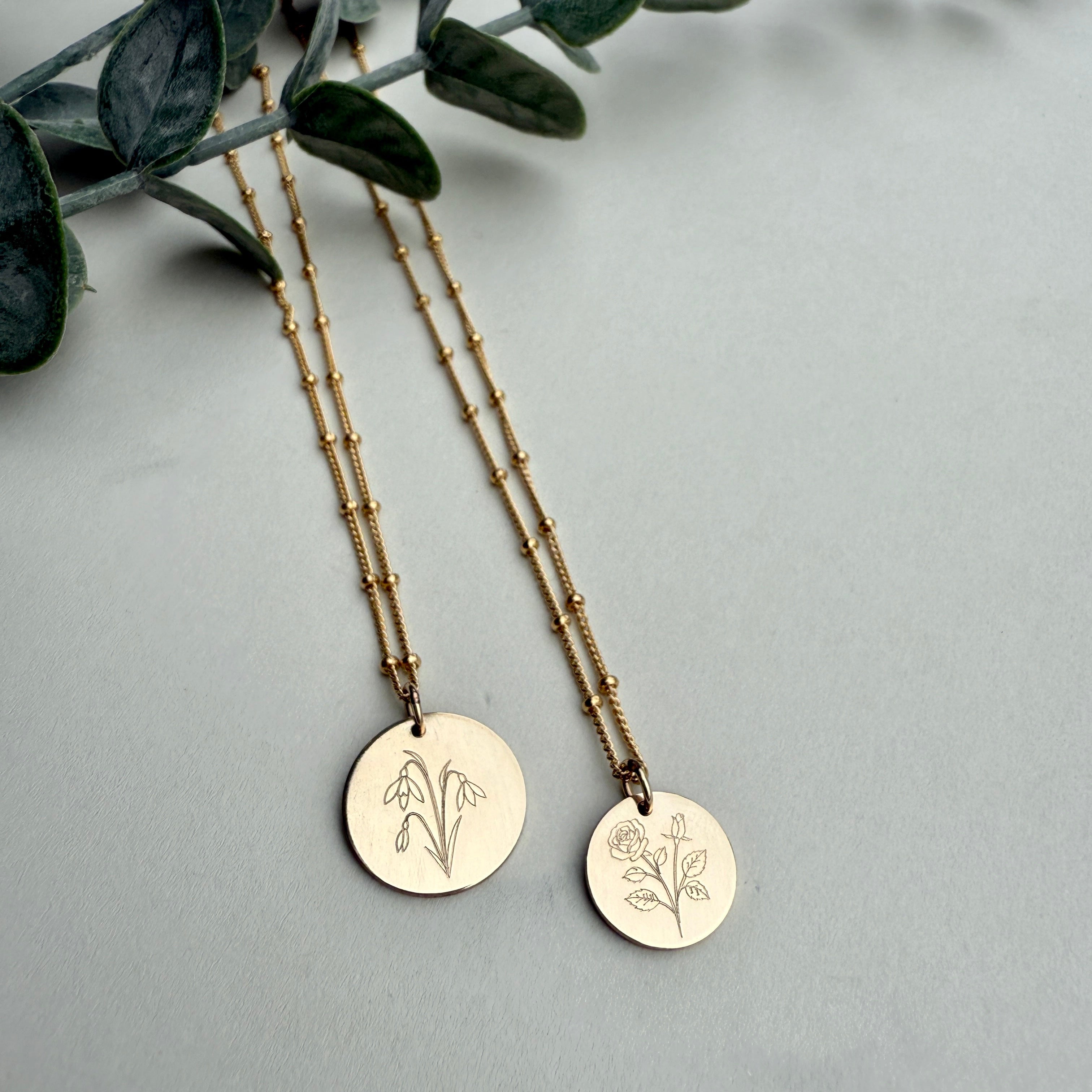 Two gold necklaces with engraved designs on a gray surface with green leaves.