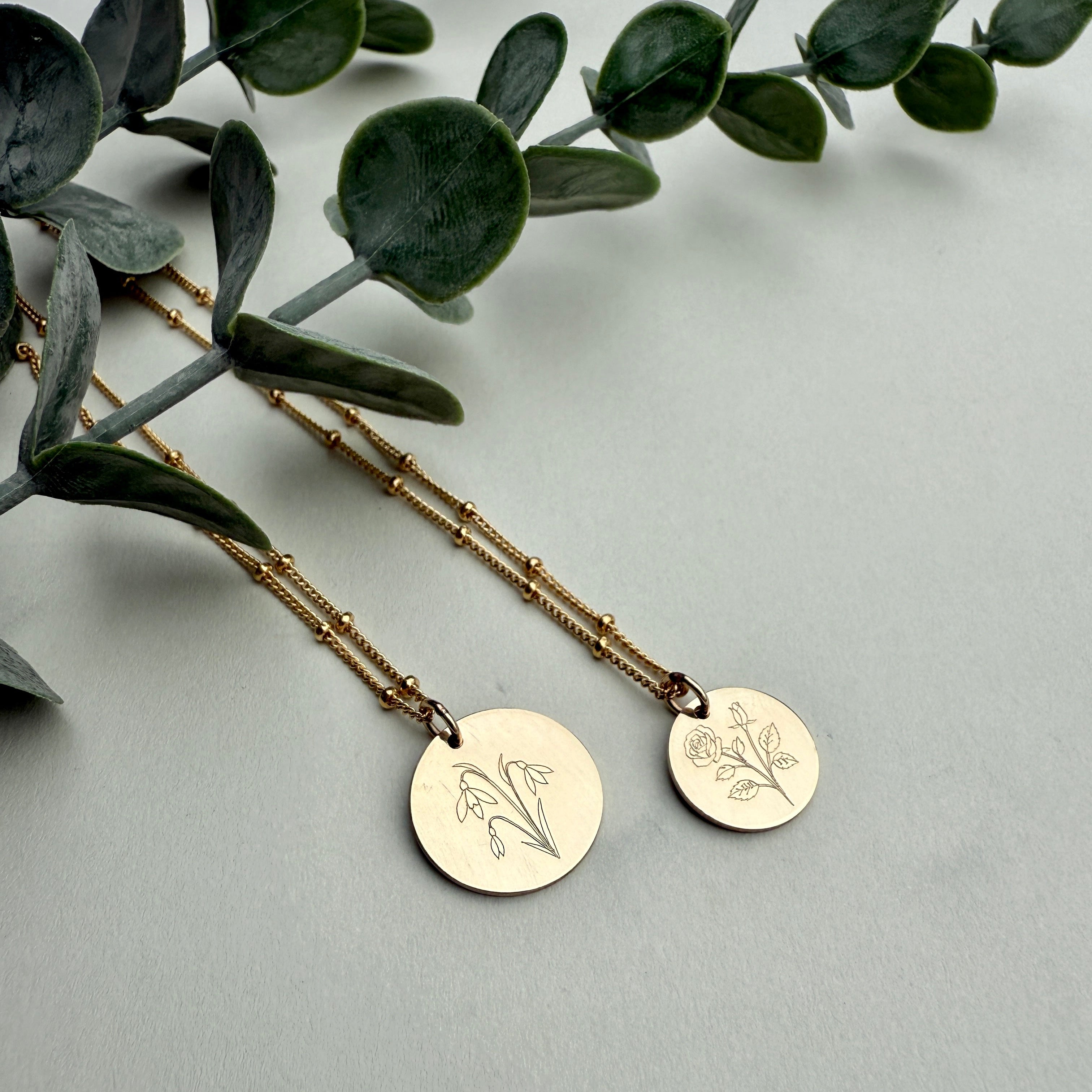 Two gold necklaces with engraved circular pendants on a light gray background with green leaves.