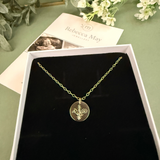 Gold filled family bouquet necklace with a pendant in a box on a green surface with flowers and a card. Flowers on pendant are snowdrop, Larkspur and cosmos