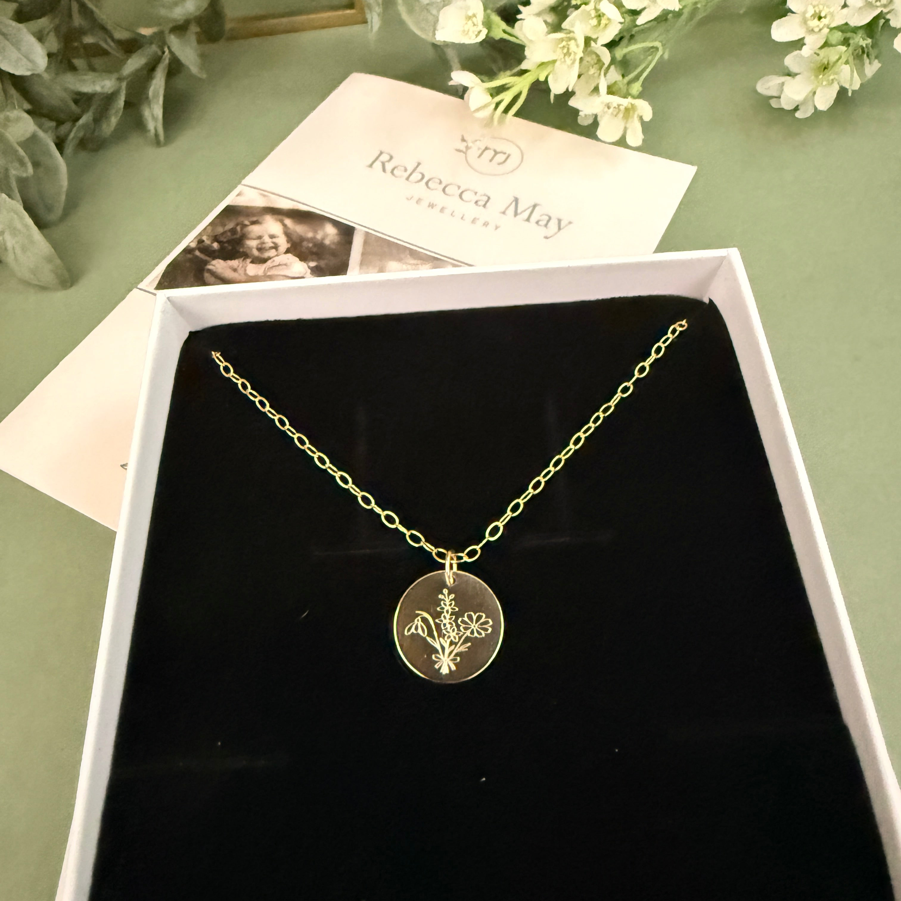 Gold filled family bouquet necklace with a pendant in a box on a green surface with flowers and a card. Flowers on pendant are snowdrop, Larkspur and cosmos
