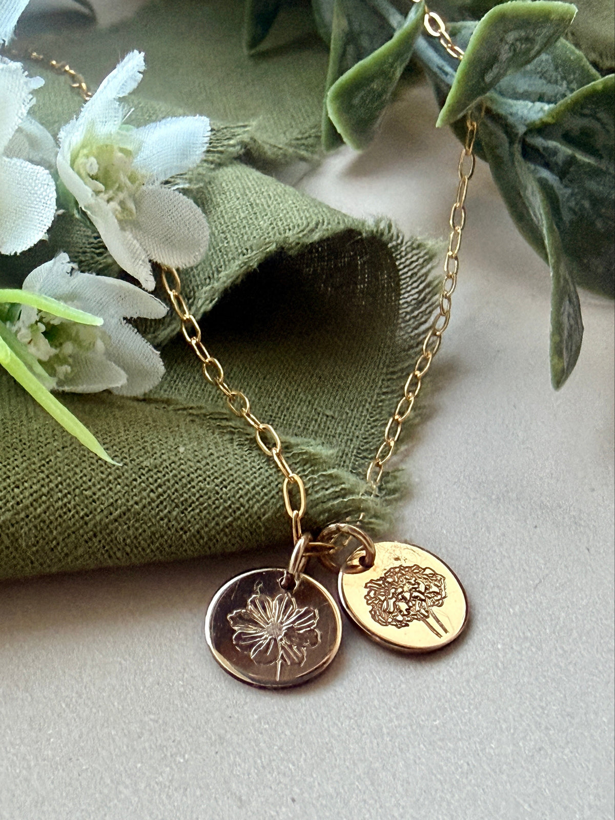 A gold chain necklace with two round engraved disc pendants, cosmos flower and marigold flower styled on an olive green fabric cloth with white flowers and green leaves.